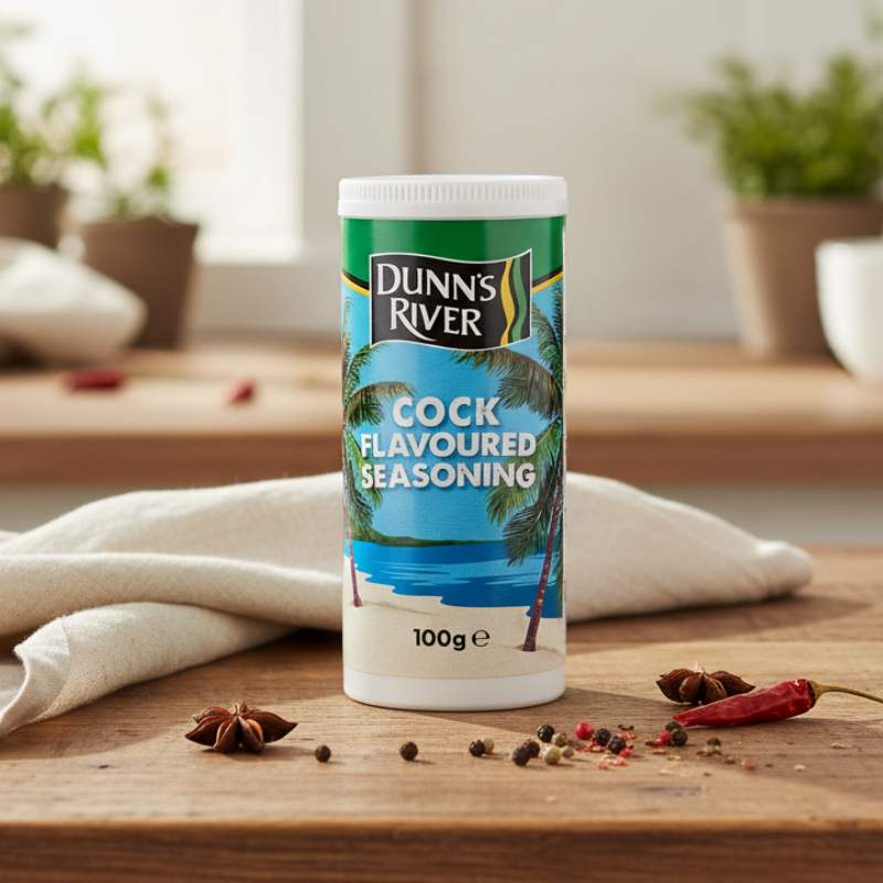 Dunns River Cock Flavoured Seasoning 100g shaker on wooden table with herbs and spices.