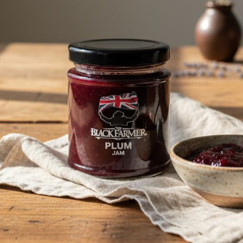 Jar of Black Farmer plum jam on a wooden table with a bowl of jam and a spoon.