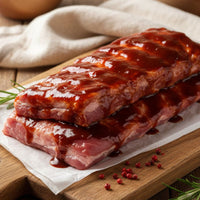 Slab of extra-meaty sticky-BBQ pork ribs on a wooden board with glaze bowl and herbs – The Black Farmer.