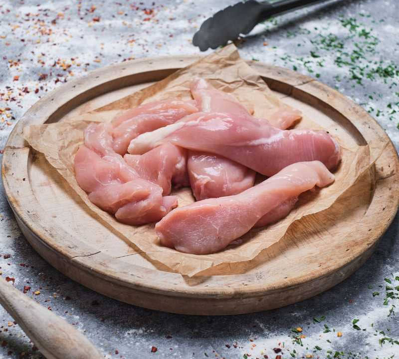 Raw chicken pieces on a wooden cutting board with spices and a wooden spoon in the background.