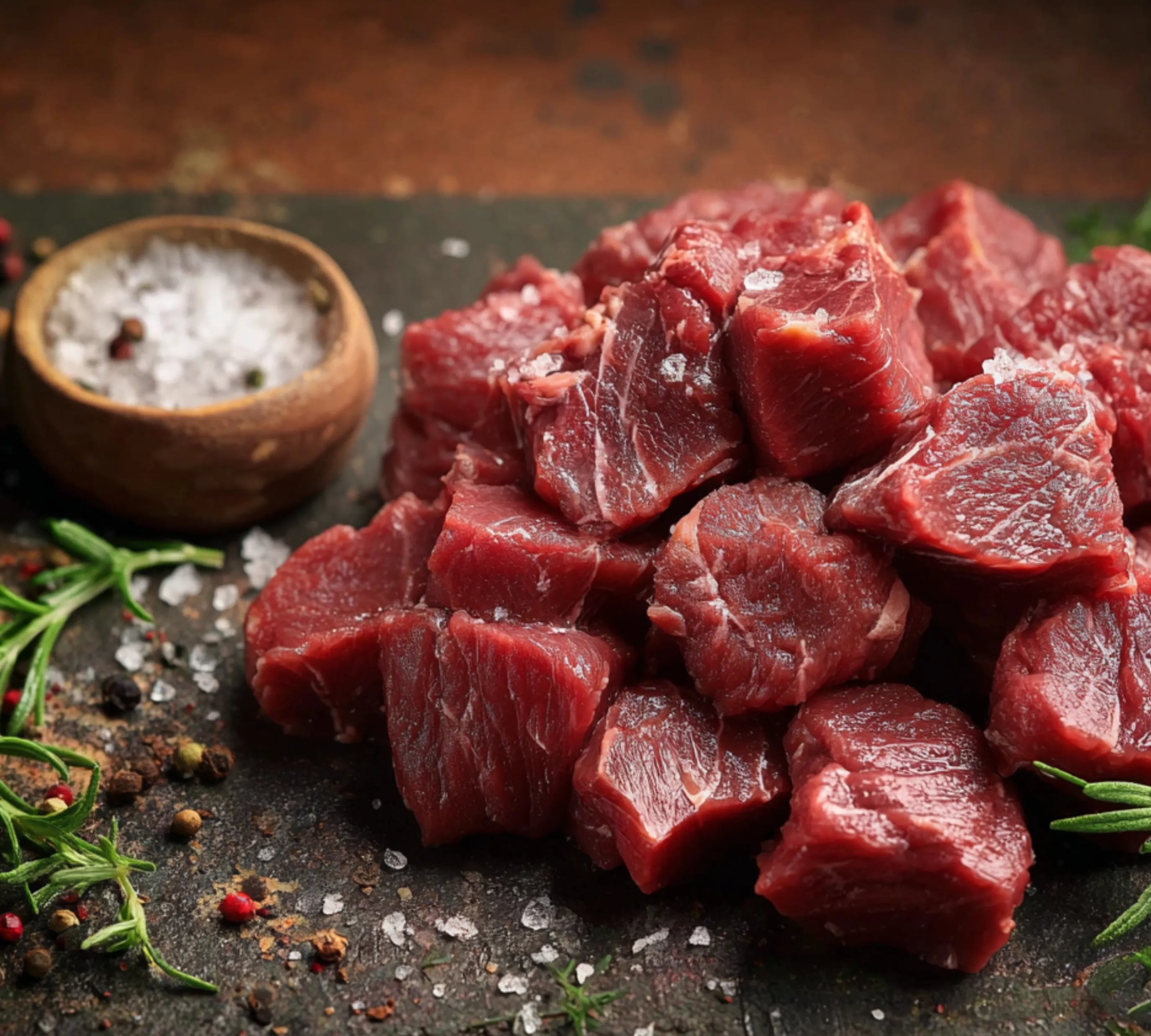 Raw beef chunks on a dark surface with herbs and spices
