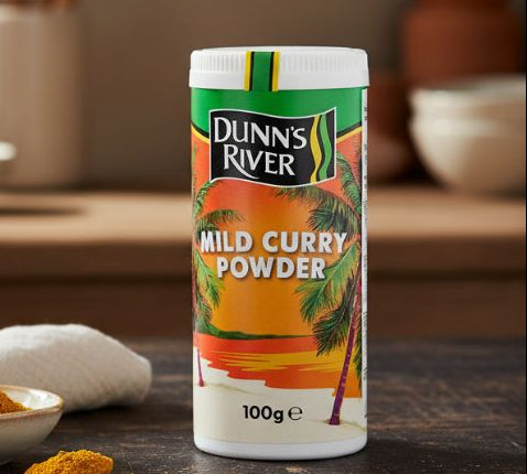 Dunns River Cock Flavoured Seasoning 100g shaker on wooden table with herbs and spices.