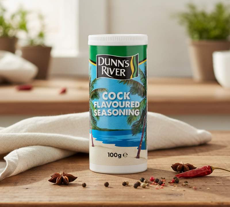 Dunns River Cock Flavoured Seasoning 100g shaker on wooden table with herbs and spices.