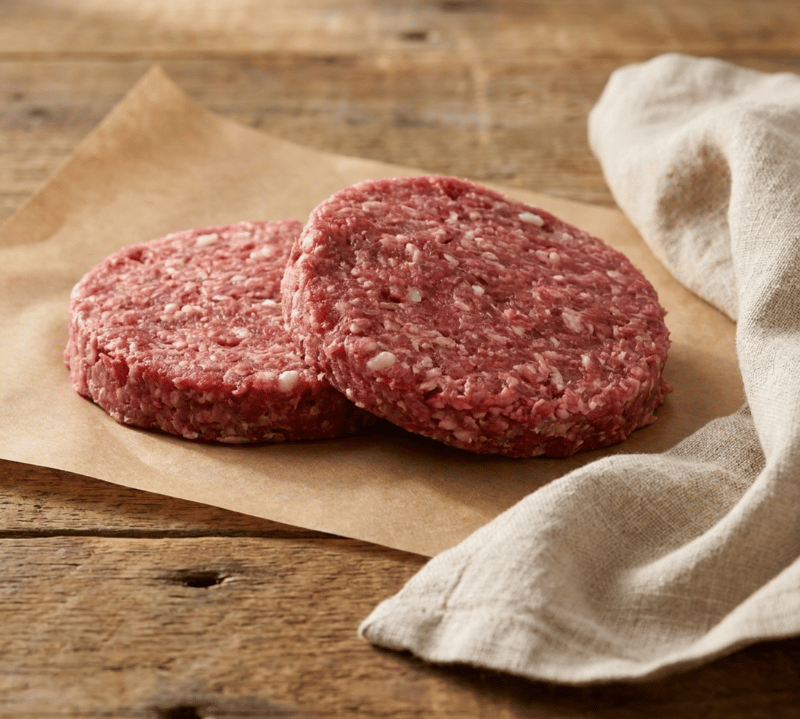 Two raw jerk-seasoned goat burger patties on a board with spices, garlic and herbs – The Black Farmer.