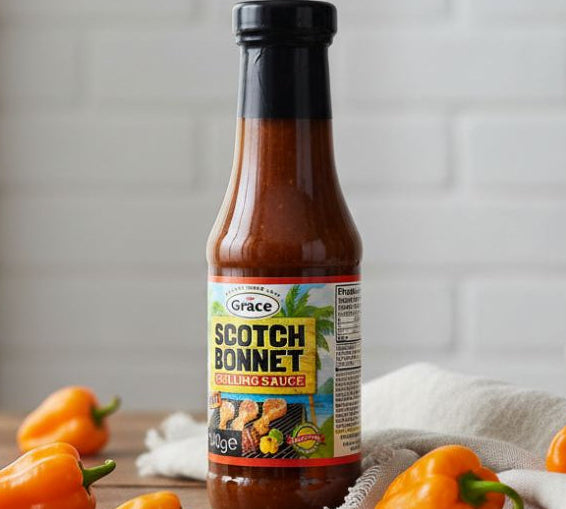 Bottle of Grace Scotch Bonnet Grilling Sauce 340g with Scotch bonnet peppers on a wooden table.