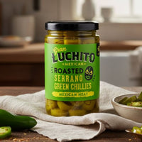 Jar of Juan Luchito roasted serrano green chillies on a wooden table with a kitchen background.