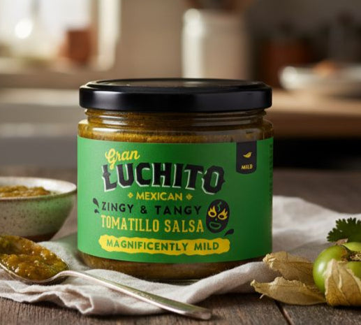 Jar of Gran Luchito Tomatillo Salsa on a wooden table with tomatoes and a bowl in a kitchen setting.