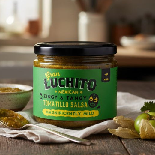 Jar of Gran Luchito Tomatillo Salsa on a wooden table with tomatoes and a bowl in a kitchen setting.
