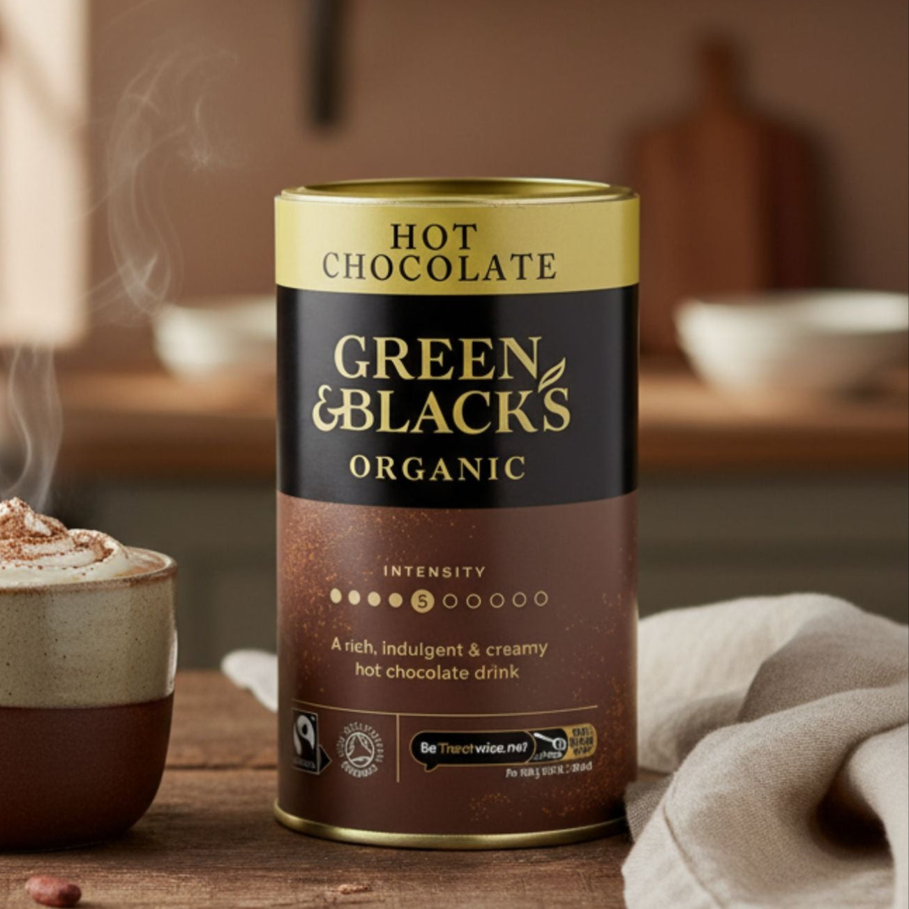 Green & Blacks Organic hot chocolate can with steaming mug on a wooden table in a kitchen setting