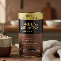 Green & Blacks Organic hot chocolate can with steaming mug on a wooden table in a kitchen setting
