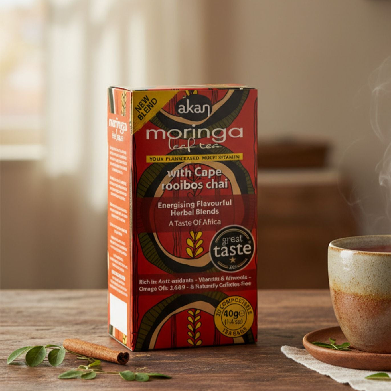 Box of Akan Moringa tea with a steaming cup on a wooden table in a cozy room.