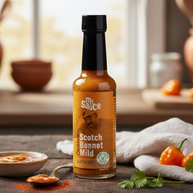 Bottle of 'My Sauce' Scotch Bonnet Mild hot sauce on a wooden surface with ingredients and a blurred background.