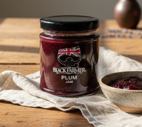 Jar of Black Farmer plum jam on a wooden table with a bowl of jam and a spoon.