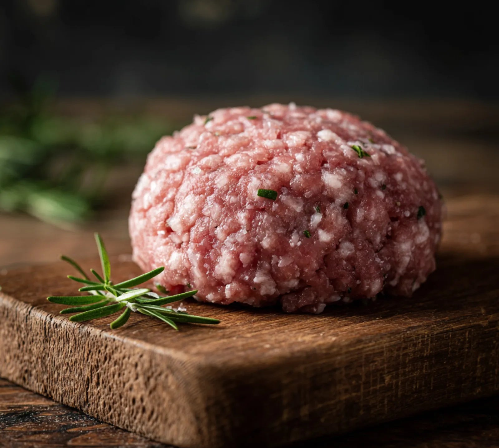 Raw ground meat on a wooden cutting board with rosemary, dark background