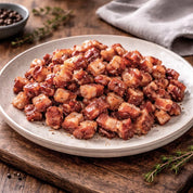 Plated dish of cooked bacon bits on a wooden surface with herbs and a towel.